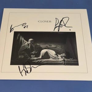 Könnte beinhalten: Ein quadratisches weißes Albumcover mit dem Text CLOSER oben und einem Schwarz-Weiß-Foto einer drapierten Figur, die auf einem Bett liegt. Drei markante Unterschriften in schwarzer Tinte sind um das zentrale Bild angeordnet.