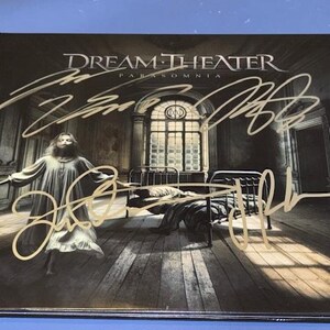 May include: Dream Theater's "Paranomnia" album with gold signatures. The cover shows a grayscale image of a woman in a flowing gown, beds, and a window. The band's name and album title are at the top.