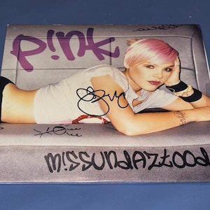 Könnte beinhalten: Ein CD-Cover mit der Künstlerin Pink, mit pinkfarbenen Haaren und einem weißen T-Shirt, liegend auf einer grauen Oberfläche. Das Wort "P!nk" steht in großen lila Buchstaben, und der Albumtitel "Missundaztood" steht unten.