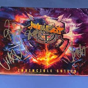 May include: A CD case with the album art for "Invincible Shield" by Judas Priest. The artwork features a shield design with a fiery background and multiple signatures in gold ink. The album title is printed at the bottom.