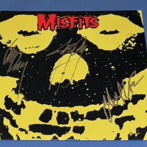 May include: A yellow and black album cover featuring the band name "Misfits" in red. The artwork includes a skull design and several signatures. The album is likely a music collectible.