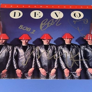 以下が含まれることがあります： デヴォの「フリーダム・オブ・チョイス」のアルバムカバー。銀色のジャンプスーツと赤い帽子をかぶった5人が写っています。アルバムタイトルは上部にあり、バンド名は大きな文字で書かれています。カバーにはバンドメンバーのサインがあります。
