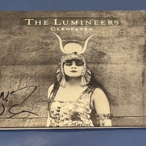 May include: A black and white album cover for The Lumineers' "Cleopatra" album. The cover features a woman in a Cleopatra-style headdress and costume. The album title and band name are printed at the top. A signature is visible.