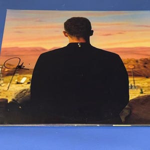 May include: A square album cover featuring a person from the back, wearing a black jacket, looking out at a desert landscape with a sunset. The album cover has an autograph in the top left corner.