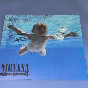 Può includere: Copertina dell'album con un bambino che nuota sott'acqua, che cerca di afferrare un biglietto da un dollaro. Il titolo dell'album "NIRVANA NEVERMIND" è stampato in nero e arancione in basso. C'è una firma nell'angolo in alto a sinistra.