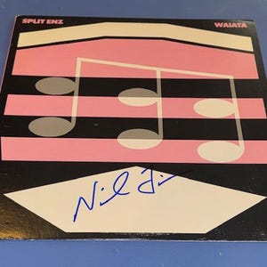 May include: A square vinyl record cover for the album Waiata by Split Enz. The cover features a black background with horizontal pink stripes and a white musical note graphic. A blue ink signature is written across the white geometric shape.