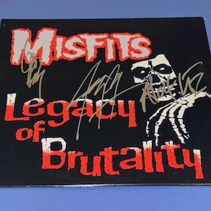 May include: Black album cover with the words "Misfits" in white and red, and "Legacy of Brutality" in red. The cover features a skeleton hand and skull graphic. Several signatures are visible on the cover.