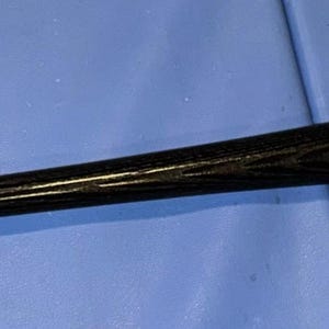Pode incluir: Um bastão de beisebol preto Louisville Slugger com uma assinatura e texto. O bastão é feito de madeira e tem um acabamento brilhante. O bastão está sobre uma superfície azul.