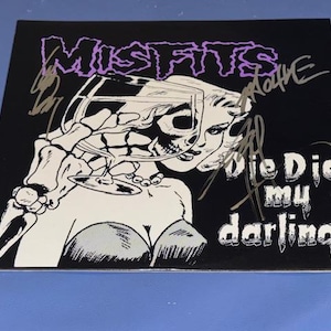 May include: Black square featuring a graphic of a skeleton holding a wine glass, with a woman's face. The word "MISFITS" is in purple, and the words "Die Die my darling" are in white. Autographs are visible.