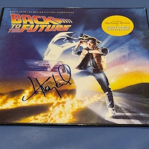 May include: A Back to the Future soundtrack album cover featuring the movie title and a graphic of the DeLorean time machine. The cover includes an image of a person and a signature. A yellow circle features the text "The Power of Love".