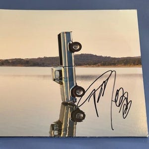 May include: A square album cover featuring a silver pickup truck standing vertically on its tailgate in the middle of a calm lake. The truck is reflected in the water below. A black ink signature is written across the lower right side of the image.