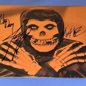 May include: Orange and black print featuring a skull-faced figure with skeletal hands, a hooded cloak, and signatures. The image has a vintage aesthetic, with the skull and hands in a lighter orange hue.