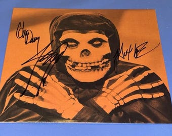 Puede incluir: Impresión naranja y negra que presenta una figura con rostro de calavera con manos esqueléticas, una capa con capucha y firmas. La imagen tiene una estética vintage, con el cráneo y las manos en un tono naranja más claro.