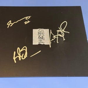 May include: A square black vinyl record sleeve featuring a central white graphic of radio waves. The cover is signed in gold ink by multiple individuals, with signatures positioned around the central graphic on the matte black surface.