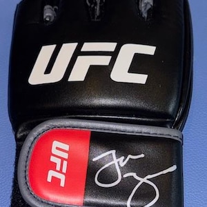 May include: Black UFC mixed martial arts glove with white UFC logo and red accent. Features a signature in white ink. The glove is designed for combat sports and training.
