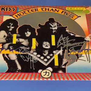 May include: A vintage KISS album cover featuring the band members in full makeup and costumes. The album title "Hotter Than Hell" is prominently displayed, with Japanese text and signatures. The cover has a red, orange, and yellow striped border.
