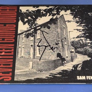 May include: Album cover for Sam Fender's "Seventeen Going Under". The image is in monochrome, showing a brick building with a signature. The album title is in red on the left, and the artist's name is in white on the bottom right.