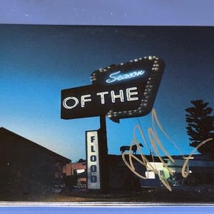 May include: A CD case with a retro neon sign that reads "Season of the Flood" against a twilight sky. The sign is black with white lettering and a blue neon outline. The case has an autograph in gold ink.