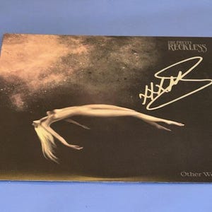 May include: Album cover for "Other Worlds" by The Pretty Reckless. The artwork features a nude figure floating in a dark, cosmic background. The band's name and album title are visible, along with a signature in white.