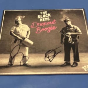 Könnte beinhalten: Werbebild für das Album "Dropout Boogie" von The Black Keys. Das Bild zeigt zwei Figuren in Arbeitskleidung, mit dem Albumtitel in rosa Schrift. Das Bild ist signiert und enthält ein Kronenlogo.