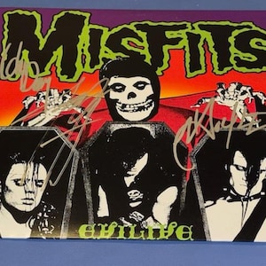 May include: A Misfits album cover with a skull and coffin design, signed by band members. The album art features a vibrant color scheme of green, orange, and purple. The word "evilive" is printed in green.