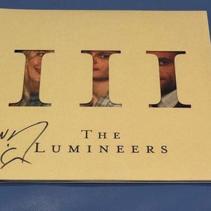 May include: A beige album cover with the word "III" cut out in three places, revealing faces. The text "THE LUMINEERS" is below the cutouts, with a signature and the initials "W.C." on the left.