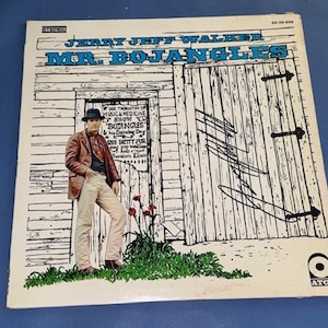 May include: A vinyl record cover for Jerry Jeff Walker's Mr. Bojangles. The cover features a line drawing of a person in a hat and jacket standing before a wooden building with a sign, accented by green grass and red flowers, with a signature.