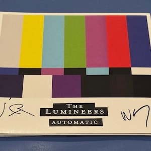 May include: A compact disc case featuring a colorful bar pattern in yellow, blue, green, purple, red, and black. The text "THE LUMEINEERS AUTOMATIC" is displayed in a black rectangle. Two signatures are visible on the case.