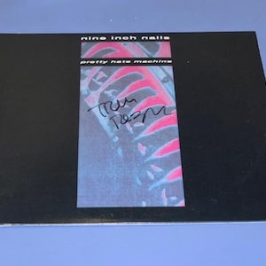 May include: A black album cover with the text "nine inch nails" and "pretty hate machine." The cover features a rectangular image with a pink and blue design and a handwritten signature.