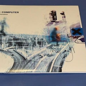 Può includere: Copertina dell'album "OK Computer" dei Radiohead. L'artwork presenta una scena autostradale in tonalità blu e bianca, con il titolo dell'album e il nome della band in alto. La copertina include anche firme manoscritte.