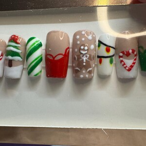 May include: A set of festive press-on nails with various Christmas-themed designs. The nails feature a mix of colors, including white, red, green, and tan. Designs include a Christmas tree, candy canes, gingerbread man, and holiday lights. The nails are arranged in a row.