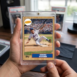May include: A baseball card of Cosmo Kramer from Seinfeld sliding into home base, encased in a protective sleeve. The Mets logo is on the player's uniform. The card has a wooden-effect border and the Topps logo is visible.