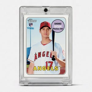 May include: A Topps Heritage baseball card featuring Shohei Ohtani in a white Los Angeles Angels jersey with a red cap, holding a black bat. The card displays the text Shohei Ohtani Pitcher/DH, Angels, and 17, encased in a clear protective plastic holder.