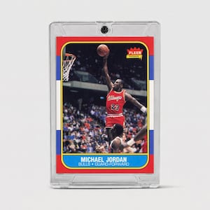 May include: A Fleer Premier basketball trading card featuring Michael Jordan in a red Chicago Bulls uniform, number 23, jumping to dunk a basketball. The card is encased in a clear protective plastic holder with red, white, and blue borders.