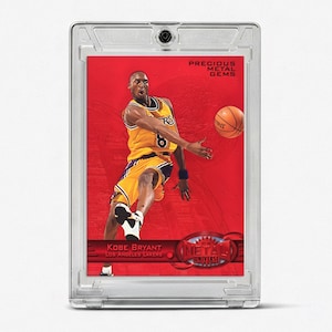 May include: A Kobe Bryant Precious Metal Gems trading card encased in a clear protective plastic holder. The card features a red background with an image of the player in a yellow Los Angeles Lakers uniform passing a basketball. Text includes Kobe Bryant, Los Angeles Lakers, Precious Metal Gems, and Metal Universe.