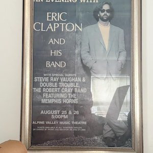 May include: Framed concert poster for "An Evening With Eric Clapton and His Band" featuring Stevie Ray Vaughan & Double Trouble, The Robert Cray Band, and the Memphis Horns. The poster is dated August 25 & 26 at 5:00 PM.