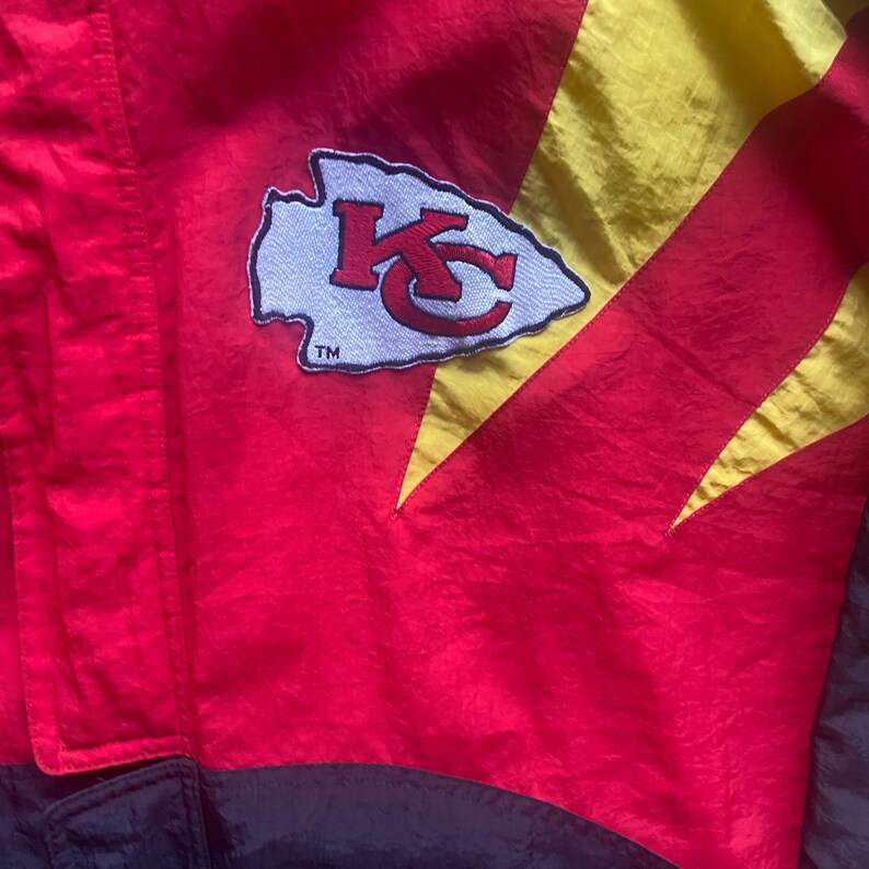 May include: A red, yellow, and black jacket with the Kansas City Chiefs logo. The logo is a white arrowhead with a red "K" and "C" inside. The jacket has a shiny, nylon-like texture.