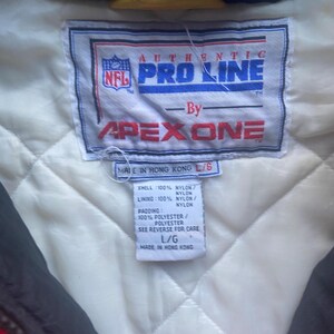 May include: Close-up of a tag from an NFL Pro Line jacket by Apex One. The tag is white with blue and red text, and includes the NFL logo. The tag also lists the materials: nylon and polyester. The jacket is a size L/G and made in Hong Kong.