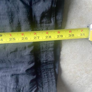 May include: A black garment is measured with a yellow tape measure. The tape measure shows measurements from 24 to 31 inches. The garment appears to be made of a shiny, possibly nylon, material.