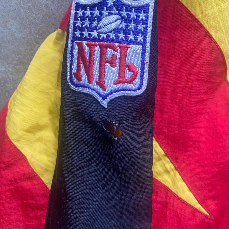 May include: Close-up of a vintage jacket featuring the NFL logo patch. The jacket has a color block design with red, yellow, and black panels. A small hole is visible on the black sleeve. The NFL logo is in blue, red, and white.