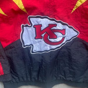 May include: A red, black, and gold jacket featuring the Kansas City Chiefs logo. The logo is a white arrowhead with a red "KC" design. The jacket has a black bottom and red top with gold accents.