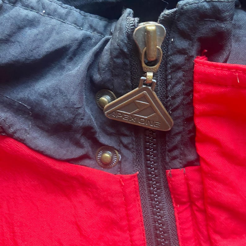 May include: Close-up of a red, black, and gray jacket. The jacket has a zipper closure with a metal Apex One tag. The jacket's hood is black, and the body is red. The zipper is closed.