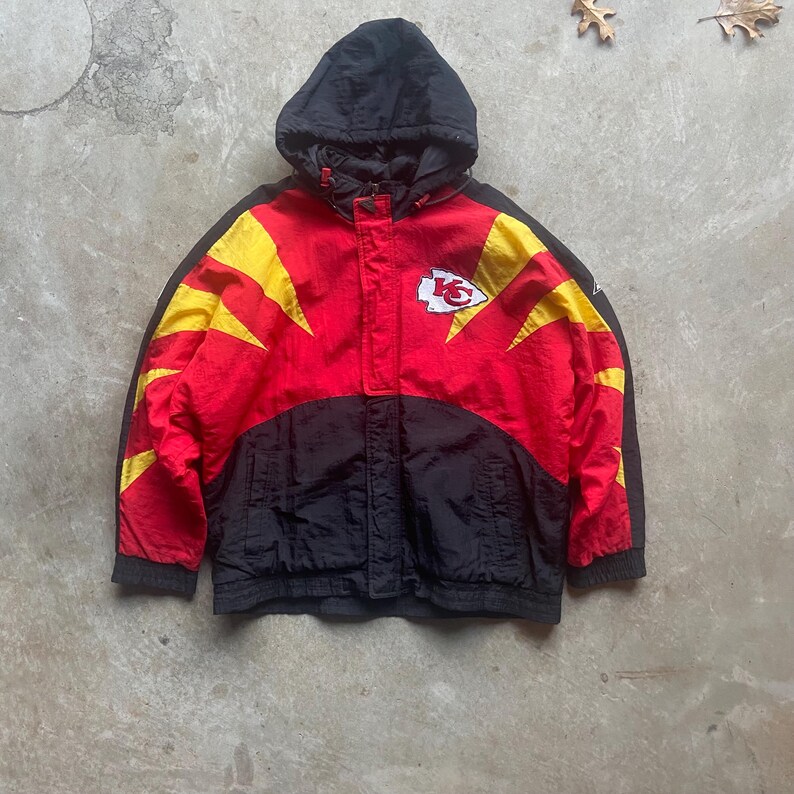 May include: A red, black, and yellow hooded jacket with the Kansas City Chiefs logo on the chest. The jacket has a black hood and sleeves, with yellow and red accents. The jacket is made of a lightweight material.