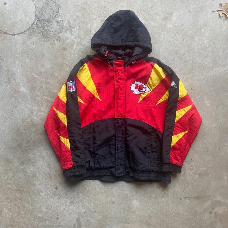 May include: A vintage hooded jacket in red, black, and yellow, featuring the Kansas City Chiefs logo and NFL patch. The jacket has a full zipper and a black hood. The design includes yellow lightning bolt accents on the sleeves and chest.
