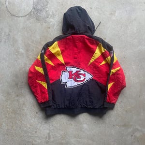 May include: A red, black, and yellow hooded jacket featuring the Kansas City Chiefs logo on the back. The jacket has a black hood and a zipper closure. The Chiefs logo is white with a red arrowhead and yellow accents.