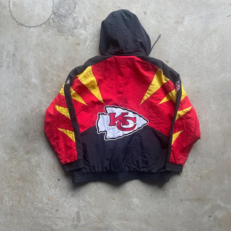 May include: A red, black, and yellow hooded jacket featuring the Kansas City Chiefs logo on the back. The jacket has a black hood and a zipper closure. The Chiefs logo is white with a red arrowhead and yellow accents.