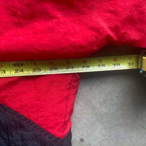 May include: A red and black jacket with a yellow measuring tape showing a length of approximately 23 to 31 inches. The jacket is made of a shiny, possibly nylon, material. The jacket is red on top and black on the bottom.