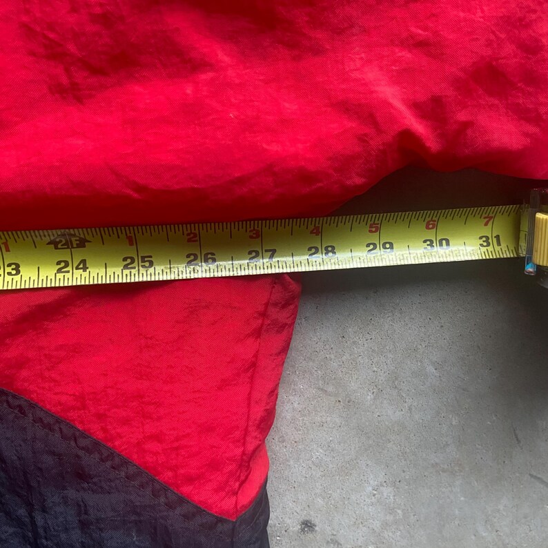 May include: A red and black jacket with a yellow measuring tape showing a length of approximately 23 to 31 inches. The jacket is made of a shiny, possibly nylon, material. The jacket is red on top and black on the bottom.