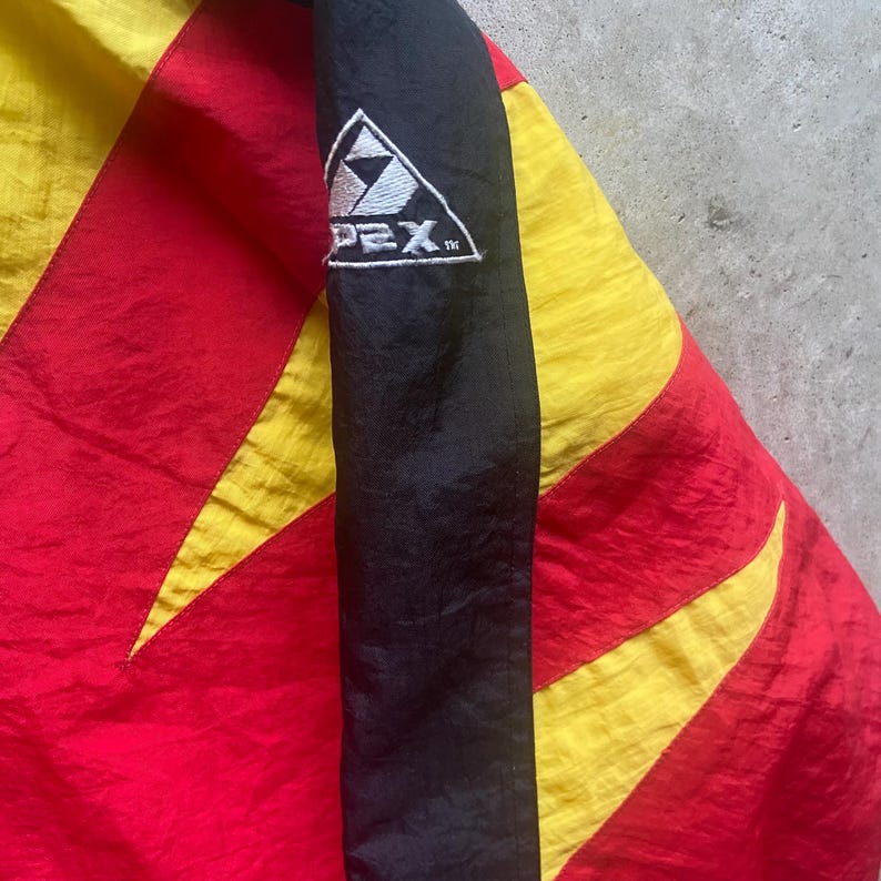 May include: Close-up of a colorful jacket with a black sleeve. The jacket features a vibrant design with red, yellow, and black panels. The sleeve displays the "PEX" logo in white within a triangle. The jacket appears to be made of a lightweight material.