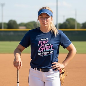 以下が含まれることがあります： ピンクと青で「I KNOW I PLAY LIKE A GIRL TRY TO KEEP UP」という文字が入ったネイビーブルーのTシャツ。青いバイザー、白いパンツ、ソフトボールグローブを着用。ソフトボールのバットが見えます。